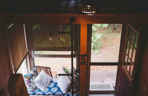Cozy interior of a tiny home with a colorful couch, large windows, and a door leading to a wooded outdoor area.