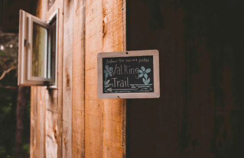 A wooden sign on a cabin wall reads "Walking Trail" with decorative flowers, next to an open window.