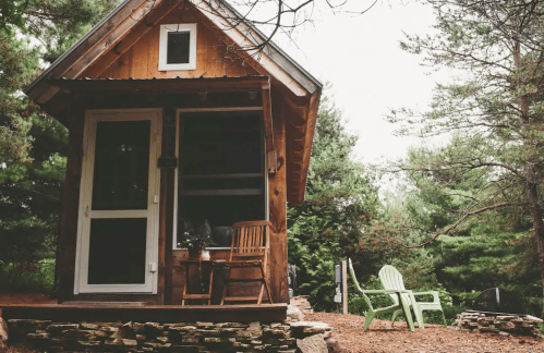 A cozy wooden cabin surrounded by trees, featuring a porch with chairs and a stone pathway.