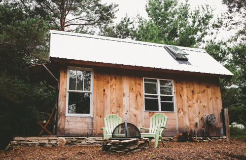 A cozy wooden cabin with a metal roof, surrounded by trees, featuring two green chairs and a fire pit in front.