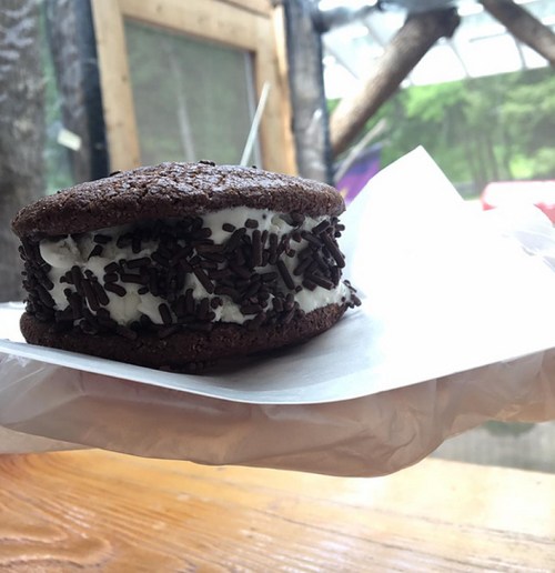 A chocolate cookie sandwich filled with white cream and topped with chocolate sprinkles, held on a paper.