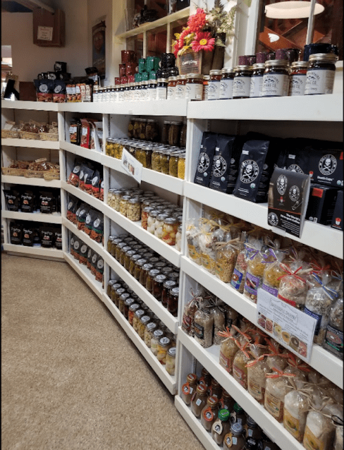 A cozy store shelf filled with jars of preserves, pickles, and various packaged goods, decorated with flowers.