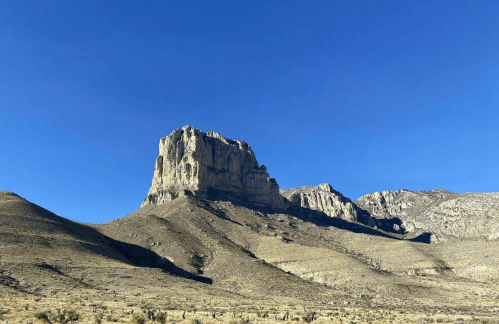 A rugged mountain peak rises against a clear blue sky, surrounded by arid terrain and rocky slopes.