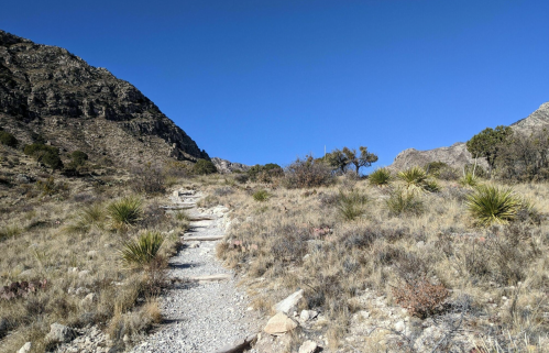 A rocky trail winds through a dry, hilly landscape with sparse vegetation under a clear blue sky.