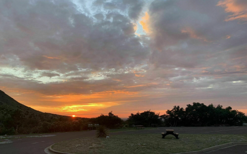 A vibrant sunset with orange and pink hues behind clouds, casting light over a grassy area and distant hills.