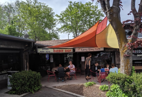 A barber shop and café with colorful awnings, outdoor seating, and people enjoying their time in a green area.