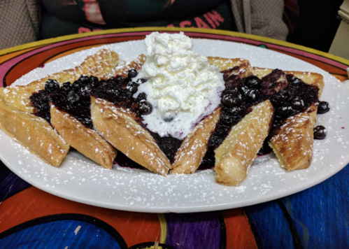 A plate of French toast topped with blueberry compote and whipped cream, dusted with powdered sugar.