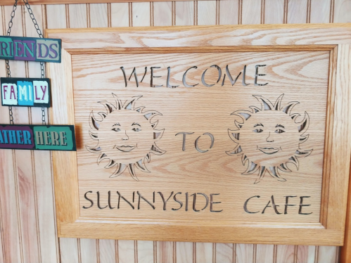 Wooden sign with carved sun faces, reading "Welcome to Sunnyside Cafe," surrounded by colorful hanging signs.