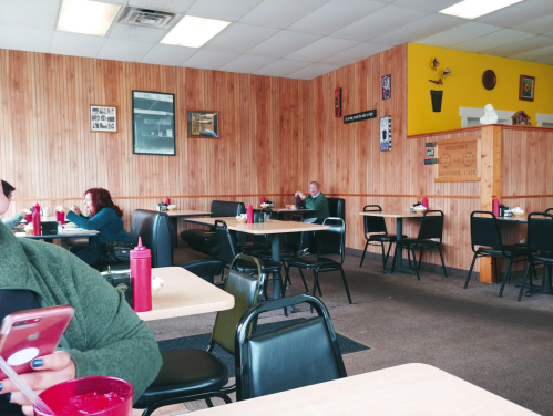 A cozy diner with wooden walls, empty tables, and a few patrons enjoying their meals. Ketchup bottles on tables.