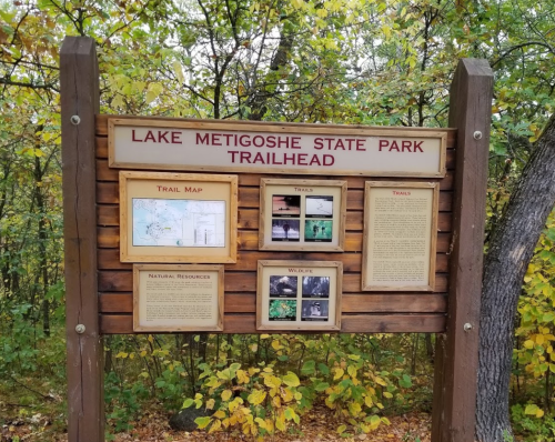 Trailhead sign for Lake Metigoshe State Park, featuring a map and information on trails and wildlife. Surrounded by autumn foliage.