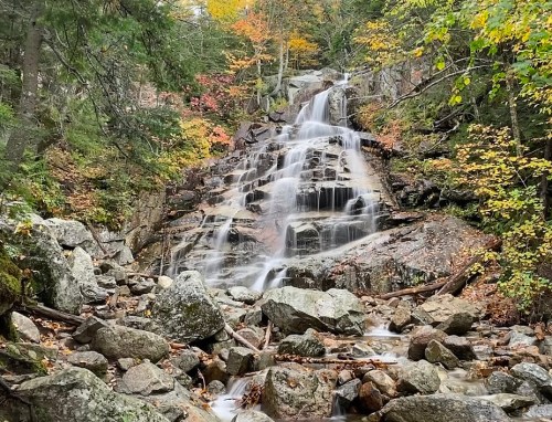 A serene waterfall cascades over rocky terrain, surrounded by autumn foliage in vibrant colors.
