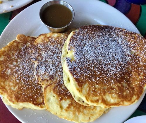 Three fluffy pancakes dusted with powdered sugar, served with a small cup of syrup on a colorful plate.