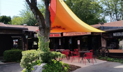A colorful orange canopy shades outdoor seating at a café, surrounded by greenery and rustic buildings.