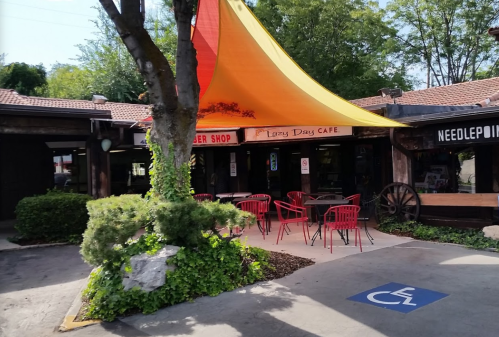 A colorful canopy shades outdoor seating at Lazy Day Café, surrounded by greenery and rustic buildings.