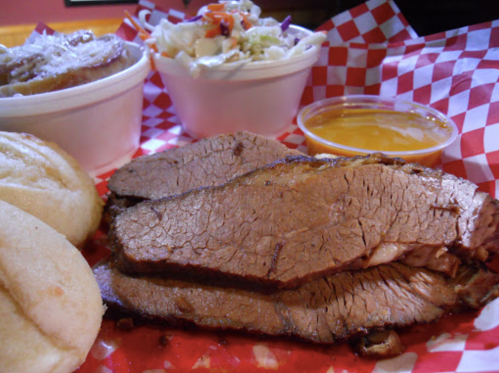 Sliced brisket with a side of coleslaw and dipping sauce, served on a red checkered tray with two bread rolls.