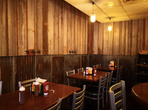 Cozy restaurant interior with wooden walls, metal accents, and tables set with condiments. Warm lighting adds ambiance.
