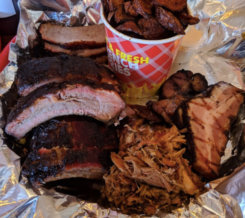 A platter of assorted smoked meats including ribs, brisket, and pulled pork, served with a cup of crispy fries.