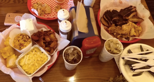A table spread with BBQ meats, sides like macaroni and cheese, cornbread, and drinks in paper cups.