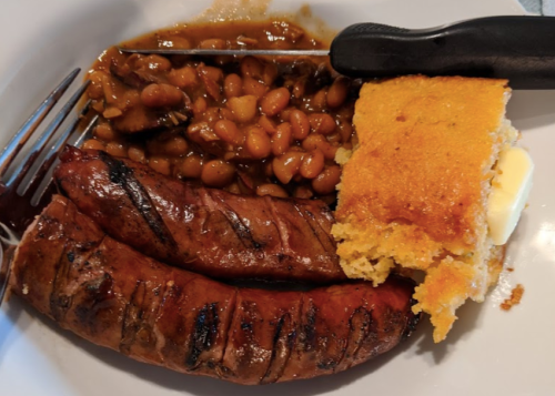 A plate with grilled sausages, baked beans, and a piece of cornbread with butter.