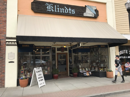 A storefront with a sign reading "Klindt's," featuring books, greeting cards, and school supplies, with a person walking by.