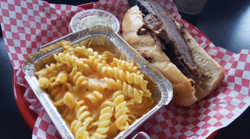 A red tray with a BBQ sandwich and a side of cheesy pasta in a foil container, garnished with pickles.