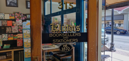 A storefront door with a sign reading "B. Lindis Booksellers and Stationers, est. 1870" and colorful book displays inside.