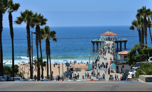 A busy beach pier with people walking, surrounded by palm trees and ocean waves under a clear blue sky.