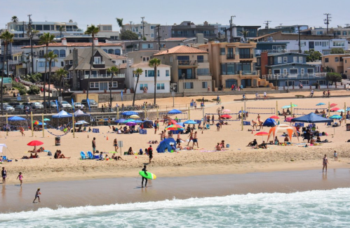 A busy beach scene with people, colorful umbrellas, and surfboards, set against a backdrop of coastal buildings.