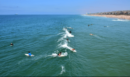 Aerial view of surfers riding waves in a clear blue ocean, with a sandy beach and city skyline in the background.