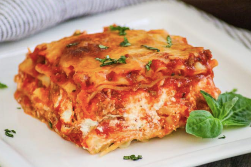 A slice of lasagna topped with herbs, served on a white plate with a striped napkin in the background.