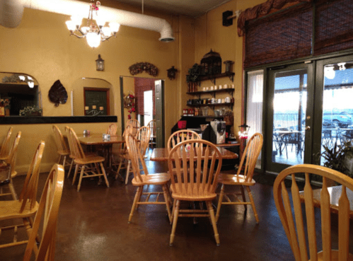 Cozy restaurant interior with wooden tables and chairs, warm lighting, and large windows overlooking an outdoor area.