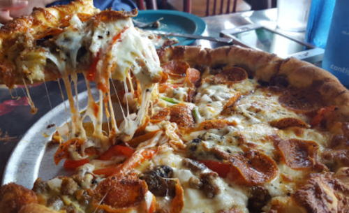 A close-up of a cheesy pizza with pepperoni, vegetables, and a hand pulling a slice away, showcasing melted cheese.