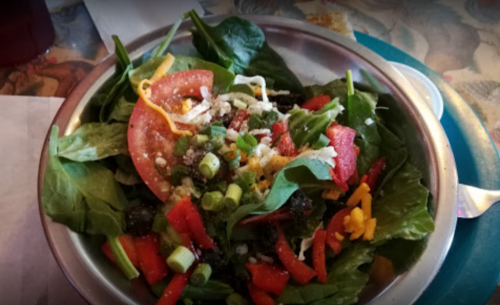 A fresh salad in a metal bowl, featuring spinach, tomatoes, green onions, cheese, and colorful vegetables.