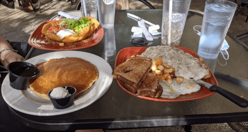 A table with a plate of pancakes, a plate of eggs with potatoes, and a sandwich, with drinks in the background.