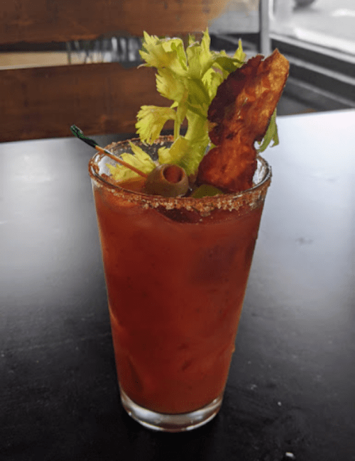 A glass of Bloody Mary garnished with celery, olives, and a crispy bacon strip, sitting on a dark table.
