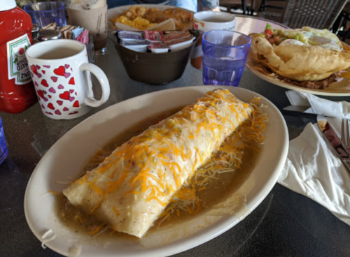 A large burrito topped with cheese and sauce, served on a plate with drinks and side dishes in the background.