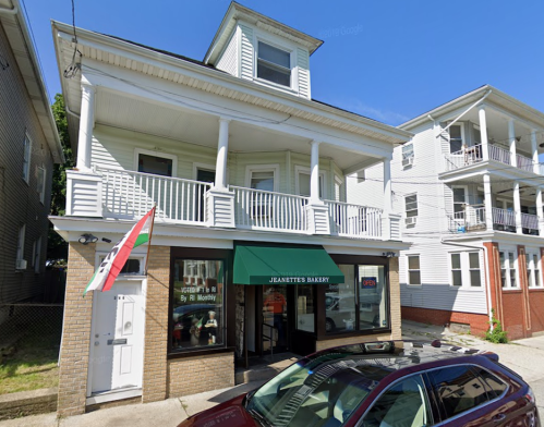 A bakery with a green awning and open sign, located in a two-story building, surrounded by residential structures.
