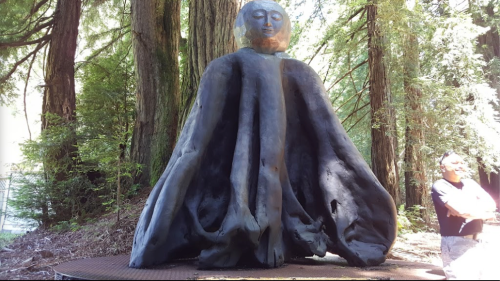 A large, abstract sculpture with a human-like figure and flowing forms, surrounded by tall trees and a person standing nearby.