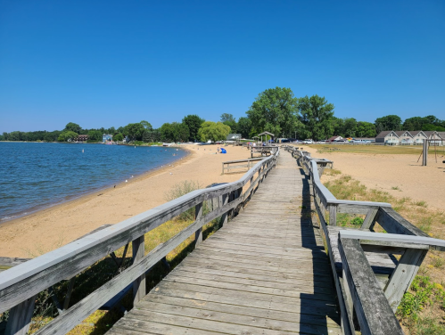 A wooden boardwalk leads to a sandy beach by a calm lake under a clear blue sky. Trees and beachgoers are visible.