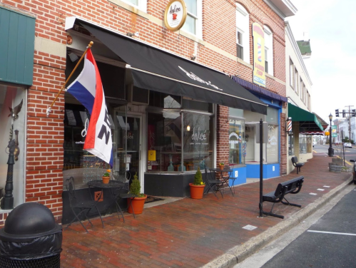 A cozy café with outdoor seating, featuring a striped awning and potted plants, located on a brick street.