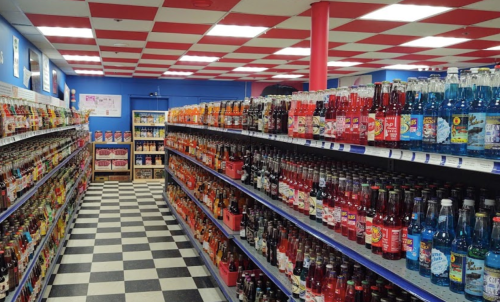 A colorful aisle in a store filled with various soda bottles on shelves, featuring a checkered floor and red and white ceiling.