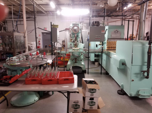A factory interior featuring machinery for bottling, with tables of empty bottles and crates in a well-lit space.