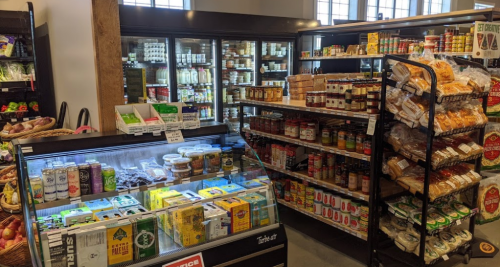 A grocery store interior featuring shelves of canned goods, snacks, and a refrigerated display case with various items.