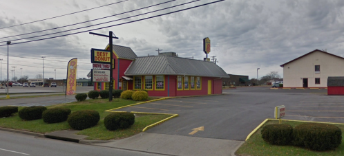 A colorful donut shop with a gray roof and signage, surrounded by a parking lot and neatly trimmed bushes.