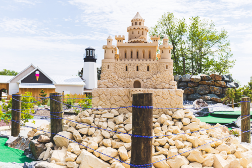A large sandcastle with towers sits atop a rocky base, surrounded by green mini-golf terrain and trees in the background.