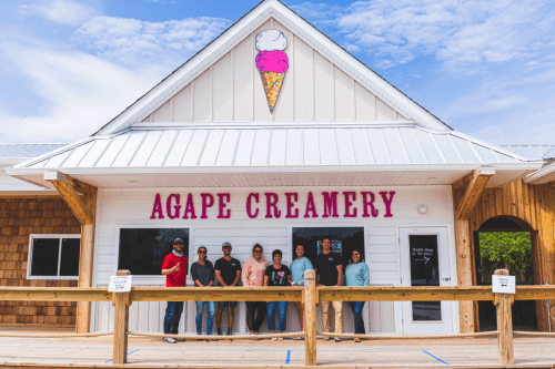 A group of people stands in front of Agape Creamery, a charming ice cream shop with a pink ice cream cone sign.