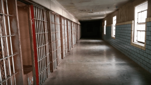 A dimly lit corridor lined with barred prison cells and concrete walls, creating a somber atmosphere.