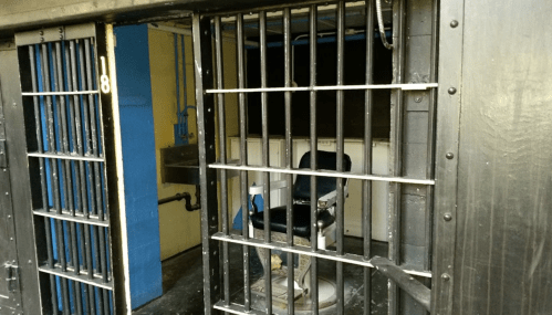 A dimly lit prison cell with barred doors and a chair inside, suggesting a solitary confinement space.