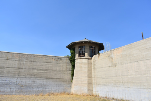A tall, concrete wall surrounds a small watchtower under a clear blue sky. Dry grass is visible in the foreground.