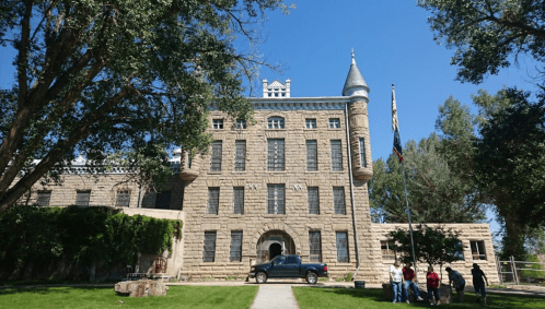 Historic stone building with a turret, surrounded by trees, and a group of people near a flagpole in front.